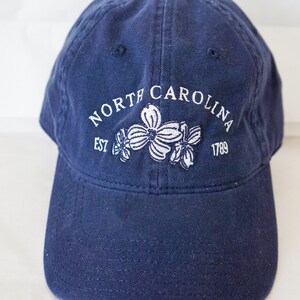 May include: A navy blue baseball cap with white embroidery that reads "NORTH CAROLINA EST 1789" and features three white dogwood flowers.