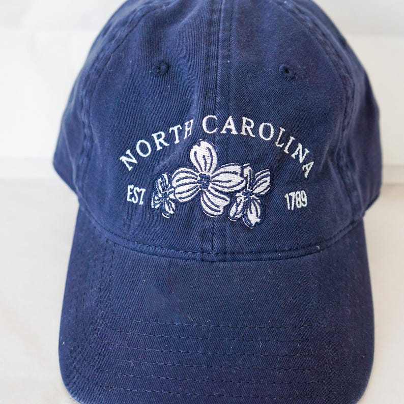 May include: A navy blue baseball cap with white embroidery that reads "NORTH CAROLINA EST 1789" and features three white dogwood flowers.