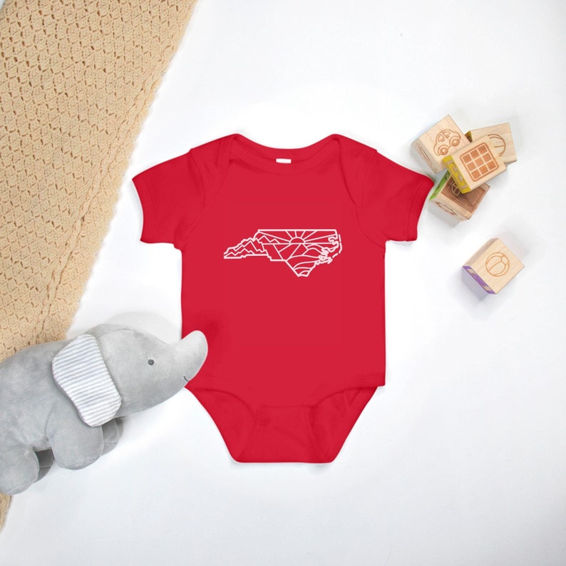 May include: Red baby onesie with a white outline of the state of North Carolina, featuring mountains and a sunrise.