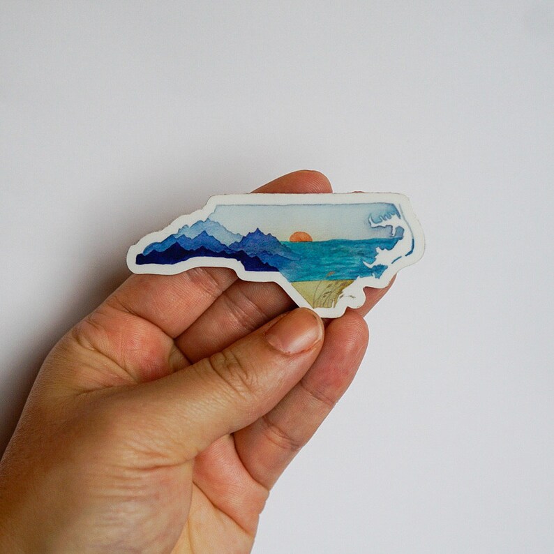 Op de afbeelding: Een sticker van de staat North Carolina met een aquarelontwerp van bergen, een zonsondergang en de oceaan.