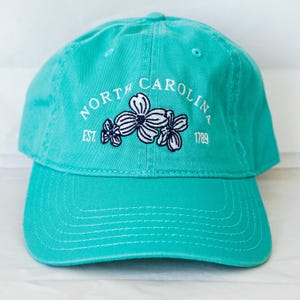 May include: A turquoise blue baseball cap with white embroidery that reads "NORTH CAROLINA EST. 1789" with a white flower design in the center.