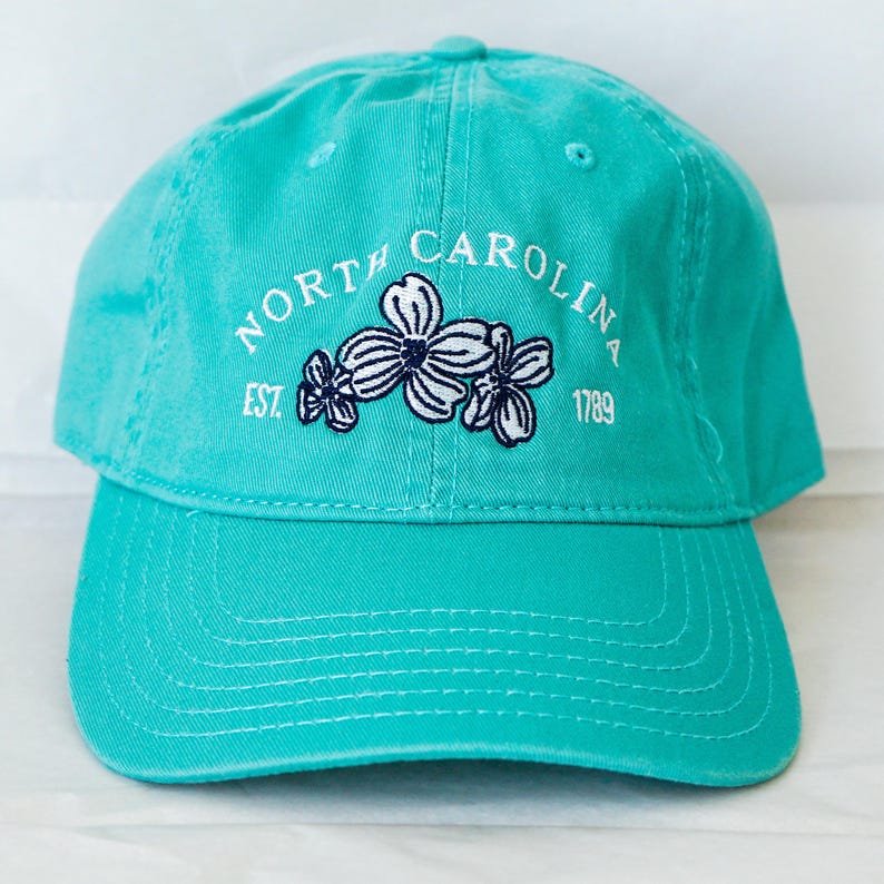 May include: A turquoise blue baseball cap with white embroidery that reads "NORTH CAROLINA EST. 1789" with a white flower design in the center.