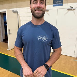 K&ouml;nnte beinhalten: Eine Person tr&auml;gt ein blaues Kurzarm-T-Shirt mit einem North Carolina-Motiv auf der Brust. Die Person h&auml;lt ein Papier und tr&auml;gt eine schwarze Smartwatch. Der Hintergrund ist eine Turnhalle.