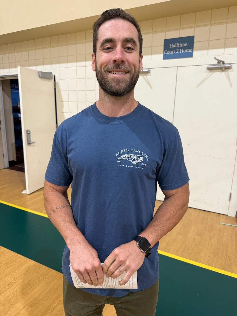 K&ouml;nnte beinhalten: Eine Person tr&auml;gt ein blaues Kurzarm-T-Shirt mit einem North Carolina-Motiv auf der Brust. Die Person h&auml;lt ein Papier und tr&auml;gt eine schwarze Smartwatch. Der Hintergrund ist eine Turnhalle.