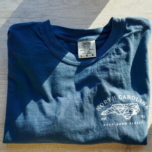 K&ouml;nnte beinhalten: Ein gefaltetes, blaues T-Shirt mit einer wei&szlig;en Grafik der Umrisse des Bundesstaates North Carolina, Bergen und Sonne. Der Text "NORTH CAROLINA" und "esse quam videri" sind ebenfalls auf dem T-Shirt aufgedruckt.