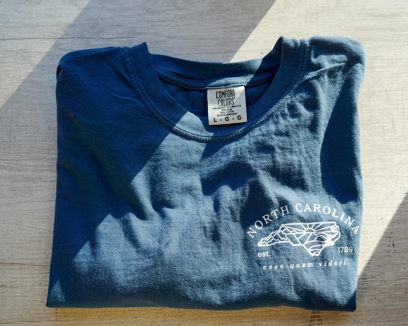 K&ouml;nnte beinhalten: Ein gefaltetes, blaues T-Shirt mit einer wei&szlig;en Grafik der Umrisse des Bundesstaates North Carolina, Bergen und Sonne. Der Text "NORTH CAROLINA" und "esse quam videri" sind ebenfalls auf dem T-Shirt aufgedruckt.