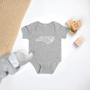 May include: A gray baby onesie with a white outline of the state of North Carolina, featuring mountains and a sun.