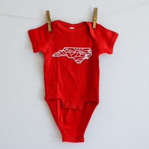 May include: Red baby onesie with a white outline of the state of North Carolina, featuring mountains and a sunrise.