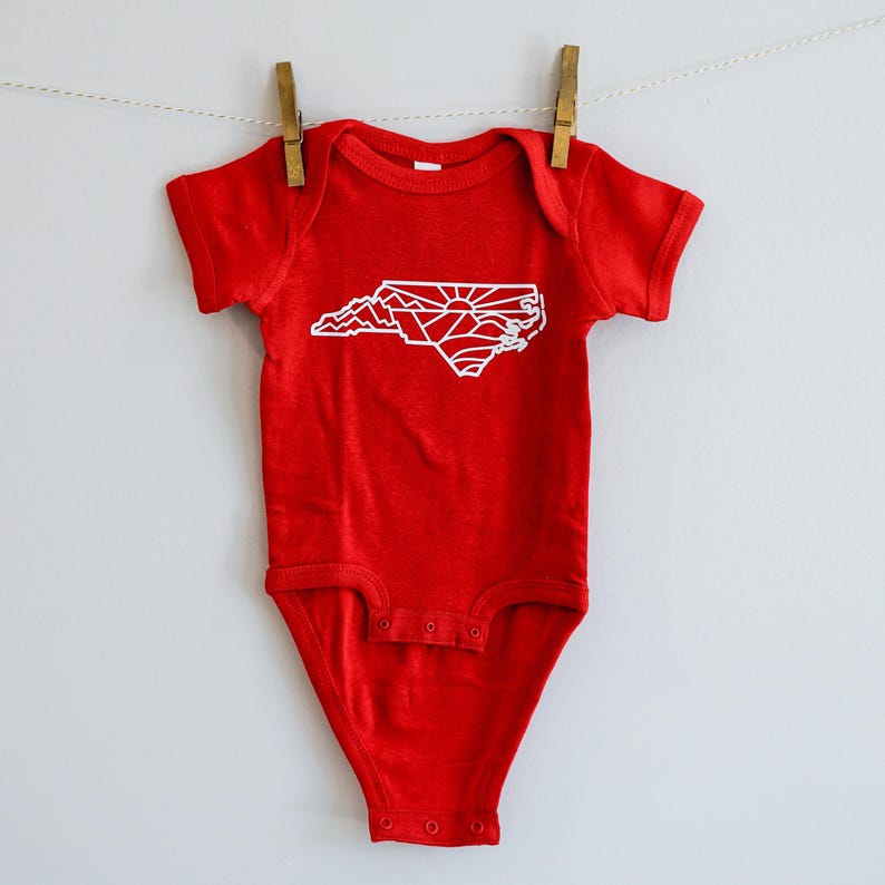 May include: Red baby onesie with a white outline of the state of North Carolina, featuring mountains and a sunrise.