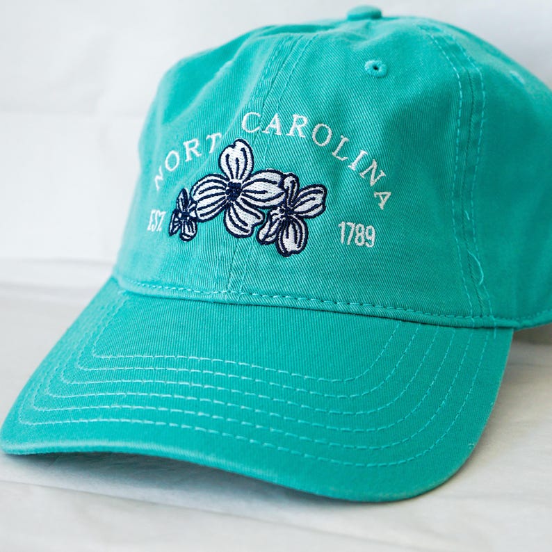 May include: A teal blue baseball cap with white embroidery that reads "NORTH CAROLINA EST 1677 1789" and features a white and navy blue embroidered flower design.
