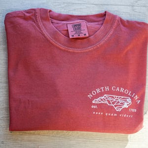 K&ouml;nnte beinhalten: Gefaltetes, ziegelrotes T-Shirt mit Rundhalsausschnitt und kurzen &Auml;rmeln. Es zeigt eine wei&szlig;e Grafik der Umrisse von North Carolina, den Text "NORTH CAROLINA", das Jahr "1789" und den Satz "esse quam videri". Das Shirt hat ein Etikett mit der Aufschrift "COMFORT COLORS".