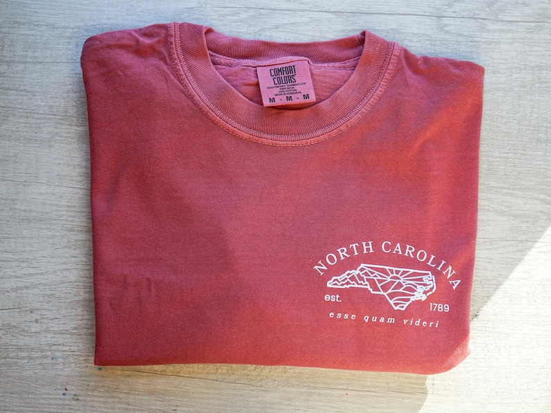 K&ouml;nnte beinhalten: Gefaltetes, ziegelrotes T-Shirt mit Rundhalsausschnitt und kurzen &Auml;rmeln. Es zeigt eine wei&szlig;e Grafik der Umrisse von North Carolina, den Text "NORTH CAROLINA", das Jahr "1789" und den Satz "esse quam videri". Das Shirt hat ein Etikett mit der Aufschrift "COMFORT COLORS".