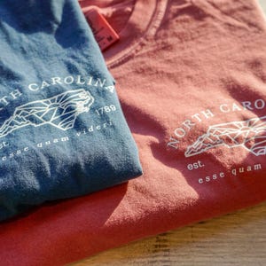 K&ouml;nnte beinhalten: Zwei gefaltete T-Shirts, eines blau und eines rot, sind gestapelt. Jedes Shirt zeigt eine wei&szlig;e Grafik der Umrisse von North Carolina, den Text "NORTH CAROLINA 1789" und den Satz "esse quam videri".