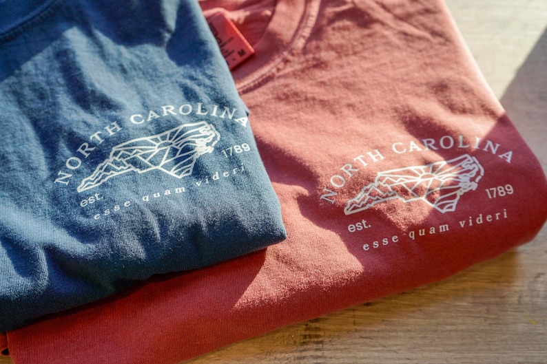 K&ouml;nnte beinhalten: Zwei gefaltete T-Shirts, eines blau und eines rot, sind gestapelt. Jedes Shirt zeigt eine wei&szlig;e Grafik der Umrisse von North Carolina, den Text "NORTH CAROLINA 1789" und den Satz "esse quam videri".