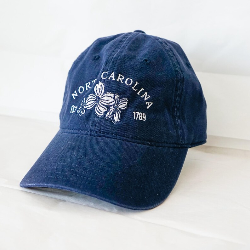 May include: A navy blue baseball cap with white embroidery that reads "North Carolina Est 1789" and features a white floral design.