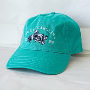 May include: A turquoise blue baseball cap with an embroidered white logo that says "North Carolina Est 1789" with a white flower design.