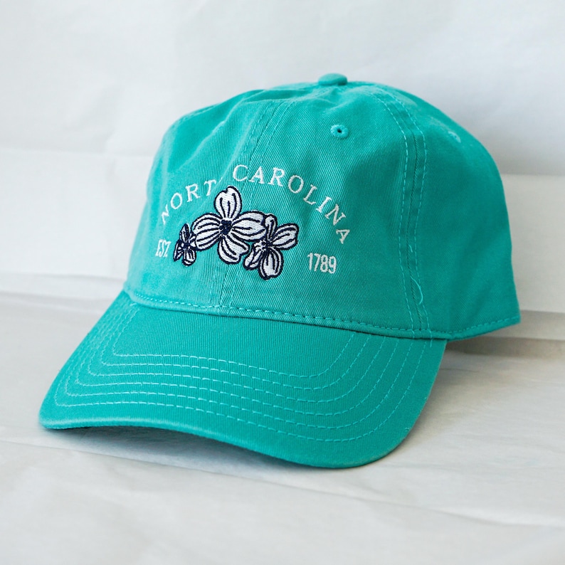 May include: A turquoise blue baseball cap with an embroidered white logo that says "North Carolina Est 1789" with a white flower design.