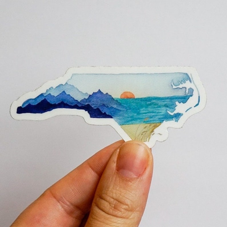 Op de afbeelding: Een aquarel sticker van de staat North Carolina met een zonsondergang boven de oceaan en bergen op de achtergrond.
