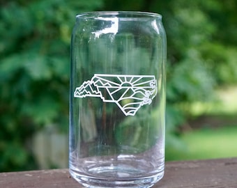 May include: A clear glass can with a white outline of the state of North Carolina, featuring mountains and a sunrise. The outline is etched into the glass.