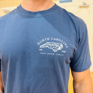 K&ouml;nnte beinhalten: Blaues T-Shirt mit wei&szlig;er Grafik der Umrisse von North Carolina, dem Text "NORTH CAROLINA" dar&uuml;ber und dem Text "est. 1789 esse quam videri" darunter. Das Shirt hat kurze &Auml;rmel und einen Rundhalsausschnitt.