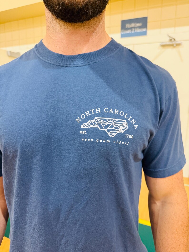 K&ouml;nnte beinhalten: Blaues T-Shirt mit wei&szlig;er Grafik der Umrisse von North Carolina, dem Text "NORTH CAROLINA" dar&uuml;ber und dem Text "est. 1789 esse quam videri" darunter. Das Shirt hat kurze &Auml;rmel und einen Rundhalsausschnitt.