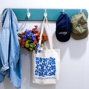 May include: A white tote bag with blue floral print and the text "NORTH CAROLINA". The bag is hanging on a teal wooden rack with a blue denim jacket, a blue baseball cap, and a green baseball cap.
