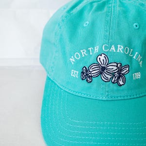 May include: A turquoise blue baseball cap with a white embroidered logo that says "NORTH CAROLINA EST. 1789" with a cluster of white flowers with navy blue centers.