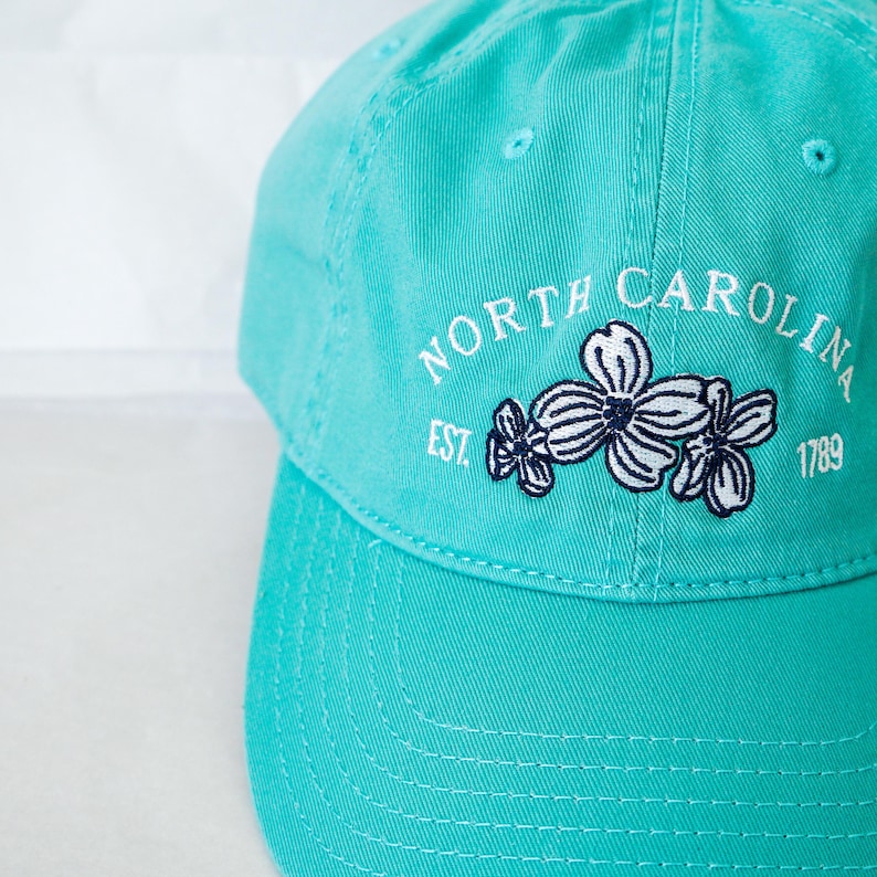 May include: A turquoise blue baseball cap with a white embroidered logo that says "NORTH CAROLINA EST. 1789" with a cluster of white flowers with navy blue centers.