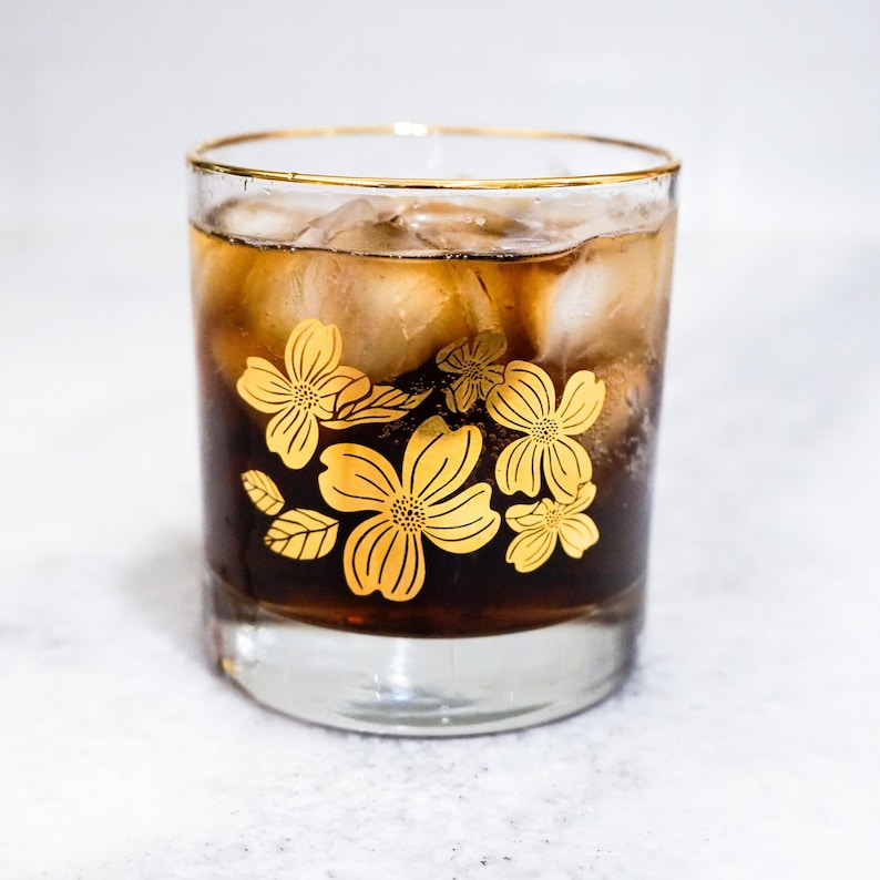 May include: A glass of cola with ice cubes in a glass with a gold rim and a floral design. The flowers are yellow and have five petals.
