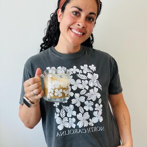 Op de afbeelding: Een vrouw draagt een grijs T-shirt met een wit bloemenpatroon en de tekst "NORTH CAROLINA" in omgekeerde volgorde. Ze houdt een glazen mok vast met een wit bloemenpatroon en een bruine drank.