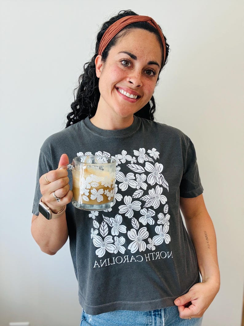 Op de afbeelding: Een vrouw draagt een grijs T-shirt met een wit bloemenpatroon en de tekst "NORTH CAROLINA" in omgekeerde volgorde. Ze houdt een glazen mok vast met een wit bloemenpatroon en een bruine drank.