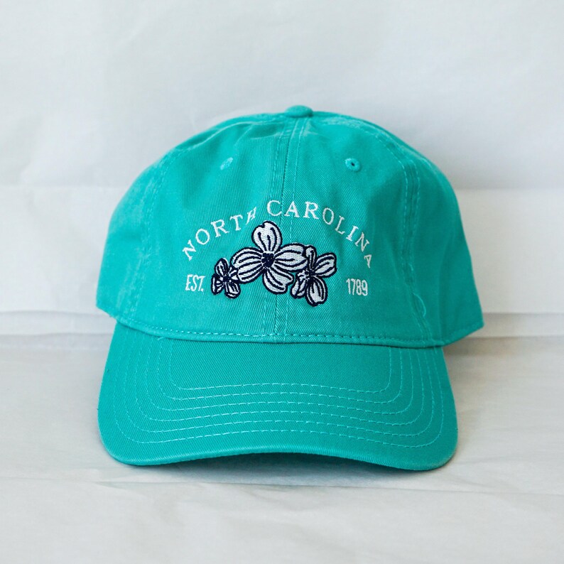 May include: A turquoise blue baseball cap with white embroidery that reads "NORTH CAROLINA EST. 1789" and features a white floral design.