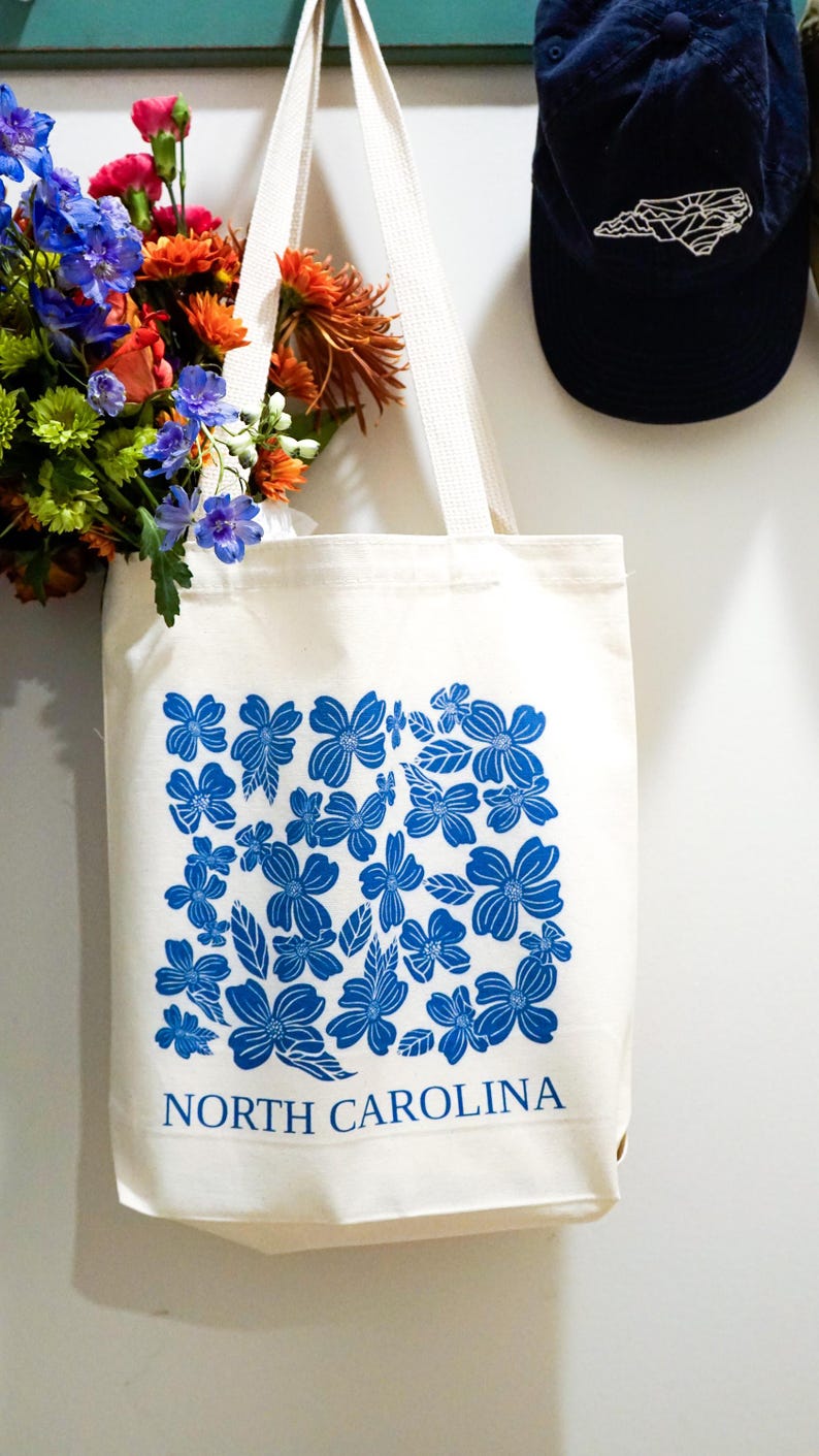 May include: A white canvas tote bag with blue floral print and the text "NORTH CAROLINA".