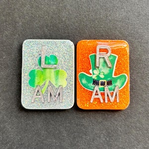 Puede incluir: Dos azulejos cuadrados, brillantes, de resina con un trébol y un sombrero de duende. El azulejo de la izquierda es plateado con un trébol verde y el texto "L AM". El azulejo de la derecha es naranja con un sombrero de duende verde y el texto "R AM".