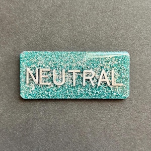 Puede incluir: Una insignia rectangular de color turquesa con purpurina plateada y la palabra "NEUTRAL" en letras mayúsculas plateadas. La insignia tiene esquinas redondeadas y está sobre un fondo gris oscuro.