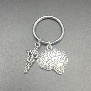 Può includere: Portachiavi color argento con un cervello e un ciondolo caduceo. Il ciondolo del cervello mostra fessure dettagliate e il caduceo ha un bastone con due serpenti e ali. L'anello del portachiavi è grande e circolare.