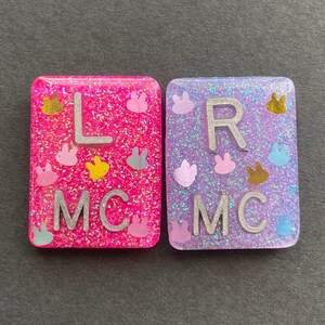 May include: Two small, square, glittery resin tiles with the letters "L MC" and "R MC" in silver lettering. The tiles are pink and purple with small, colorful bunny shapes.