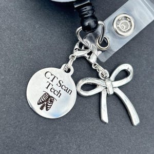 May include: A white and black retractable badge reel with the text "Have The Day You Deserve" and a skeleton graphic. It has a silver charm with a bow and a CT Scan Tech charm.