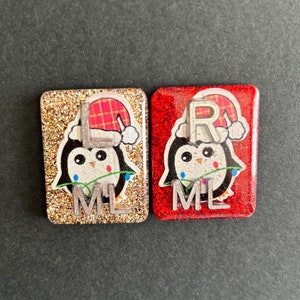 Puede incluir: Dos pequeños adornos cuadrados y brillantes con ilustraciones de pingüinos. El adorno de la izquierda es dorado con un pingüino que lleva un gorro de Papá Noel rojo y blanco y el texto "ML". El adorno de la derecha es rojo con un pingüino que lleva un gorro de Papá Noel rojo y blanco y el texto "R ML".