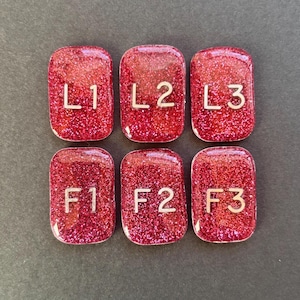 May include: Six rectangular, glittery red resin tiles. Each tile has a rounded top and contains a gold letter and number combination: L1, L2, L3, F1, F2, and F3. The tiles are arranged in two rows of three on a dark gray surface.