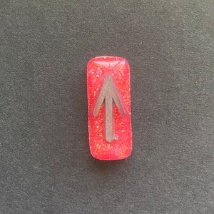 Puede incluir: Un pequeño amuleto de resina rojo lleno de purpurina con una flecha plateada apuntando hacia arriba.
