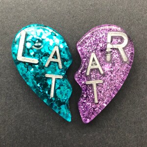 May include: Two heart-shaped pieces of jewelry with glitter, one blue and one purple, that fit together to form a whole heart. The words "LAT" and "ART" are written on the pieces.