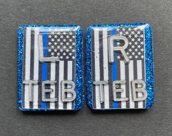 May include: Two rectangular, glittery blue accessories. Each features a black and white American flag design with a blue stripe, and the letters "L TEB" and "R TEB" in silver. The background is a dark gray.