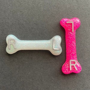 Puede incluir: Dos abalorios con forma de hueso con purpurina. Uno es blanco con letras plateadas 'R' y 'L' y el otro es rosa con letras plateadas 'T' y 'R'.