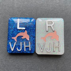 May include: Two rectangular resin pieces. One is blue with glitter, the letter "L", a pink dolphin, and the letters "VJH". The other is silver with glitter, the letter "R", a pink dolphin, and the letters "VJH".