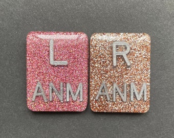 May include: Two square tiles with glitter. The left tile is pink with the letters "L ANM" in silver. The right tile is gold with the letters "R ANM" in silver.