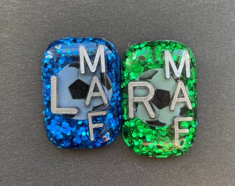 May include: Two square-shaped resin keychains with glitter. One is blue with a silver soccer ball and the letters "MARF". The other is green with a silver soccer ball and the letters "MARF".
