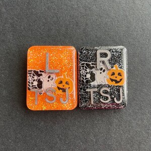 以下が含まれることがあります： ハロウィーンをテーマにした画像が描かれた、小さく正方形のキラキラ光るレジンタイルが2つ。左側のタイルはオレンジ色で、幽霊とカボチャ、そして「L」と「TSJ」の文字が描かれている。右側のタイルは黒色で、幽霊とカボチャ、そして「R」と「TSJ」の文字が描かれている。