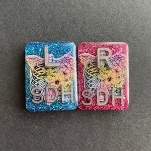 May include: Two square resin tiles with glitter embedded in the resin. The tiles are decorated with a floral and ribcage design. The left tile is blue and says "L SDH". The right tile is pink and says "R SDH".