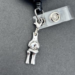 May include: A white and black badge reel with the text "The Day YOU DESERVE". A silver-coloured charm in the shape of a human elbow joint hangs from the reel. The reel has a clear plastic clip.